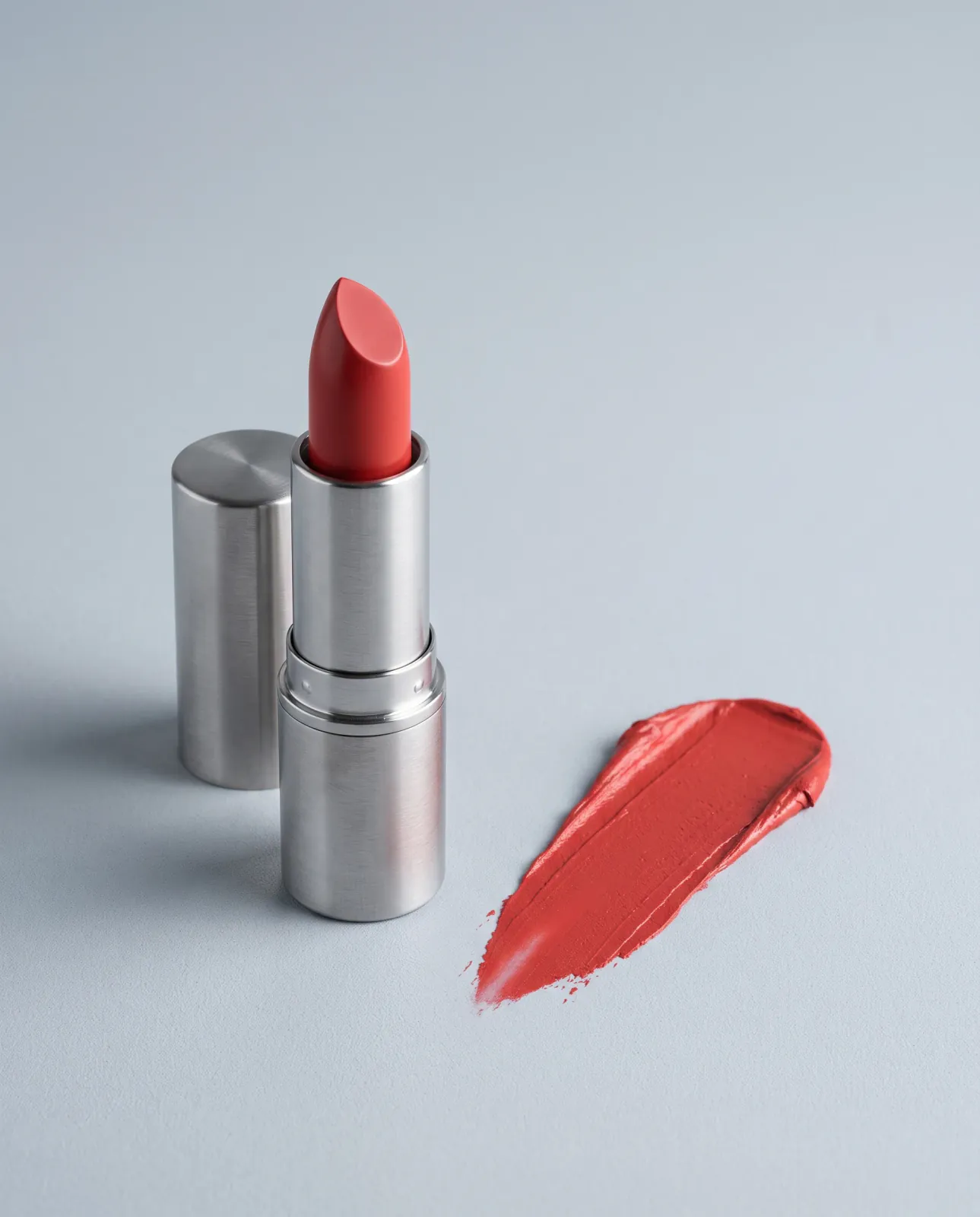 An open coral lipstick in a brushed-silver case beside its cap and a matching color swatch on a cool-gray surface.