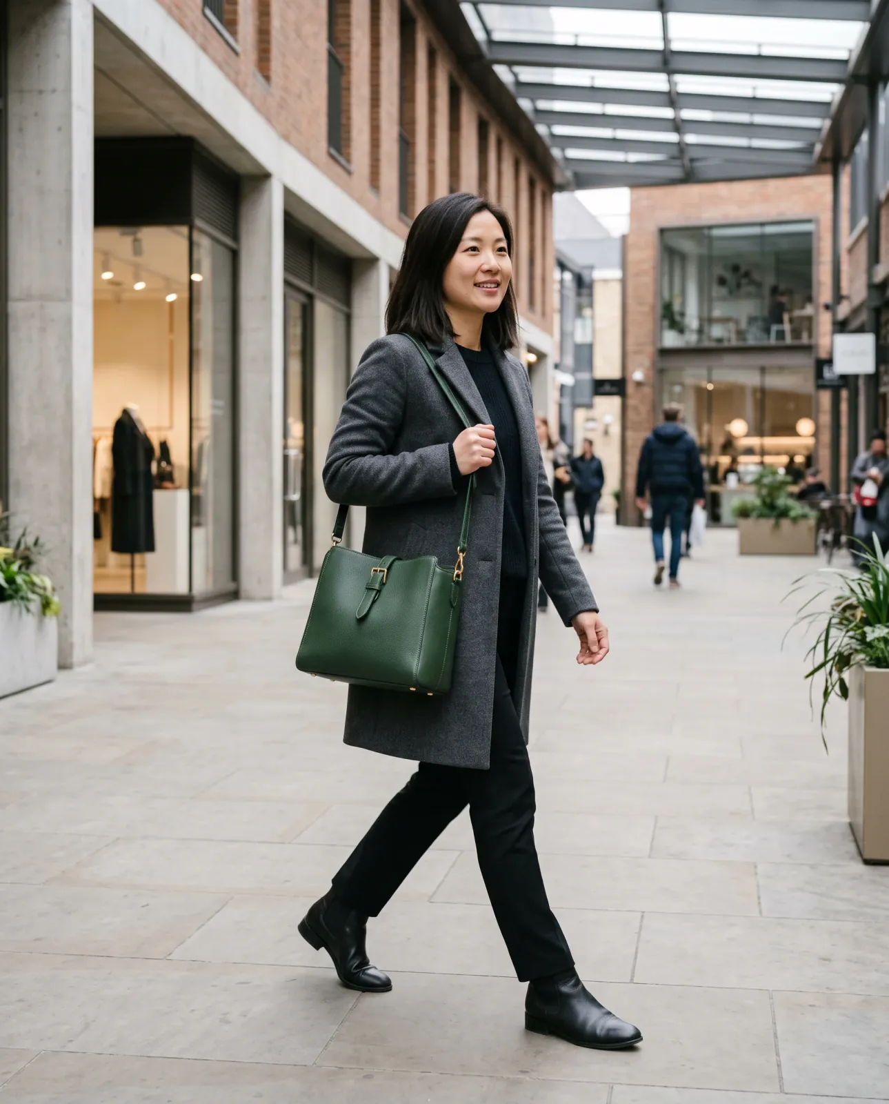 A woman in a charcoal coat carries a structured forest-green leather shoulder bag while walking through a modern shopping arcade.