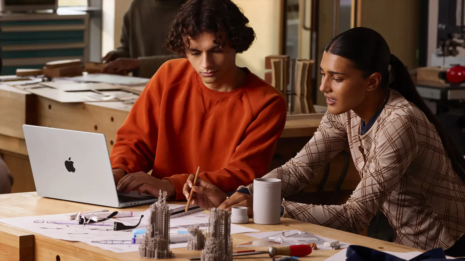 Two people use a silver 14-inch Apple MacBook Pro among sketches and architectural models.